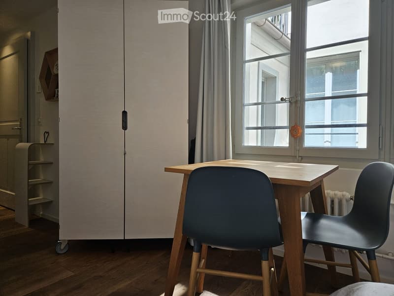 A modern, minimalist room with a wooden table, two chairs, a tall cabinet, and a small shelf. The room has large windows with grey curtains and a wooden floor.