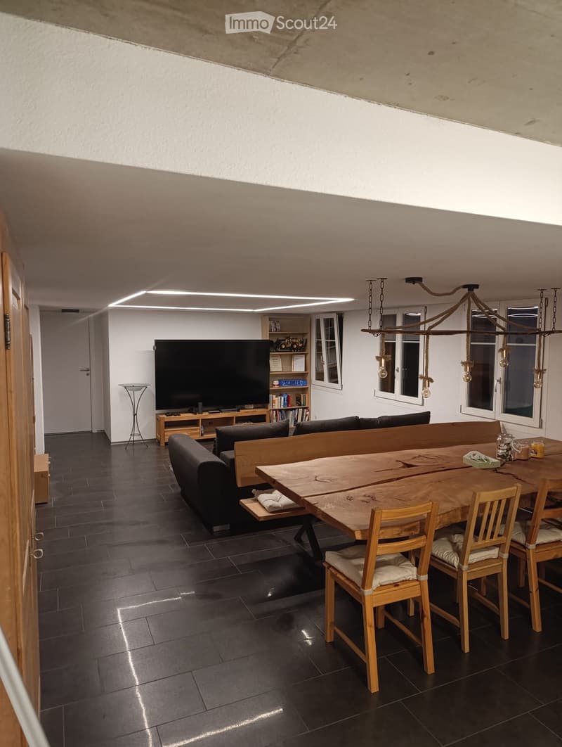 Modern living and dining area with dark tiled flooring, a large wooden dining table with chairs, a sofa, a television, a bookshelf, and a floor lamp.