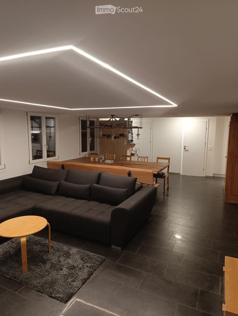 Modern living and dining area with dark tiled flooring, a sectional sofa, a small round wooden stool, a dining table with chairs, and a hanging light fixture.