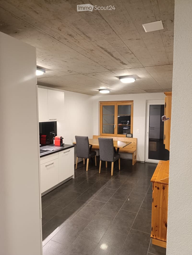 Open-plan kitchen and dining area with white cabinets, a coffee maker on the counter, a dining table with chairs, and a large window with wooden frame.