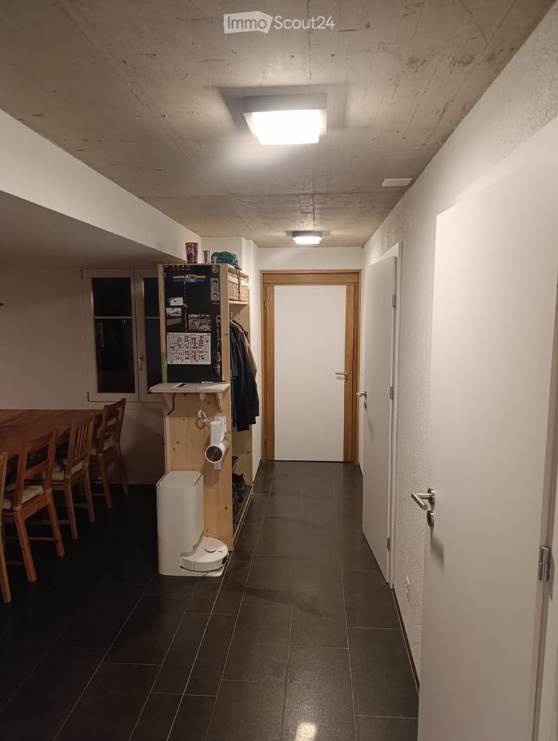 A modern hallway with dark tiled flooring, white walls, and recessed lighting. Features a small wooden shelf with a vacuum cleaner, a few chairs, and a door leading to another room.