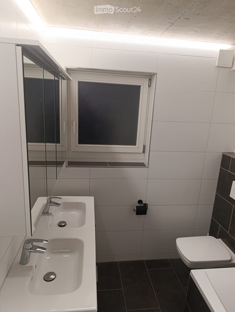 Modern bathroom with double sink, chrome faucets, toilet, and large mirror. White tiles and dark floor.