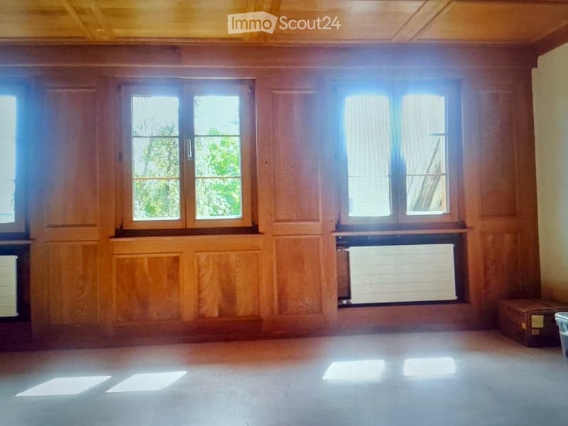 Empty room with wooden panel walls, large windows, and a radiator beneath one window. The floor is light-colored and the room is well-lit with natural light.