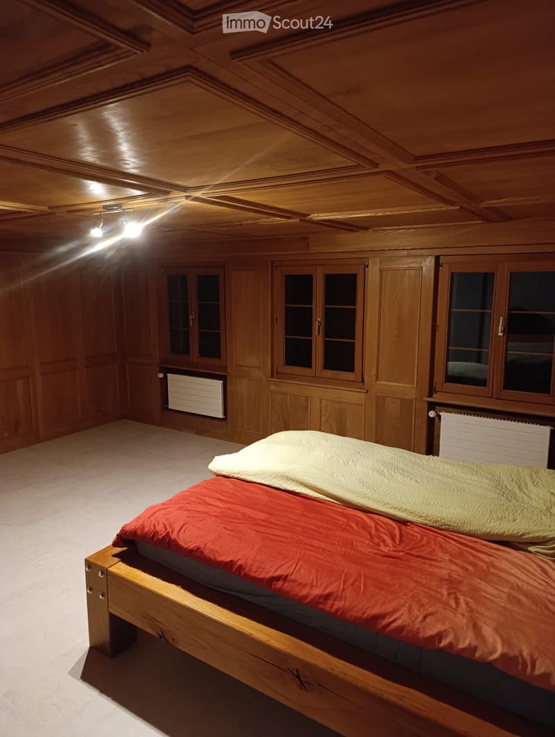 Cozy wooden bedroom with a single bed, red and yellow blankets, wooden walls, and built-in wooden cabinets with glass doors.