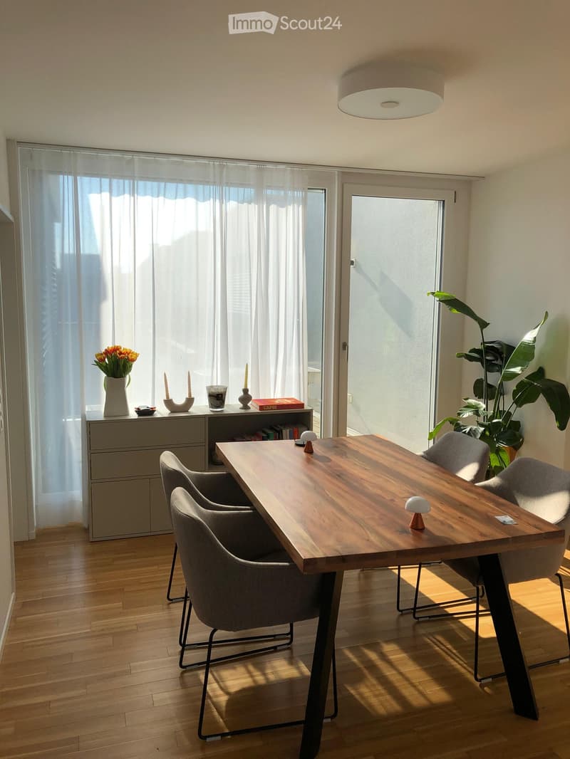 Bright dining area with a wooden table, four grey chairs, a grey cabinet, a vase with flowers, candles, and a large plant near the window.