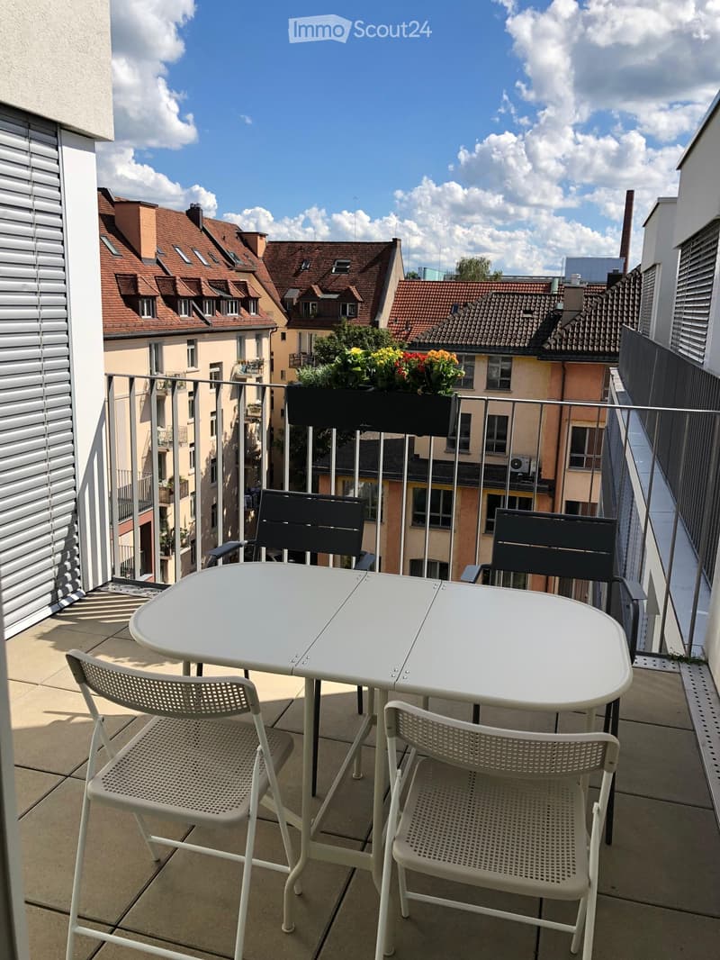 Balcony with a white table and four chairs, overlooking a cityscape with red-roofed buildings and greenery.