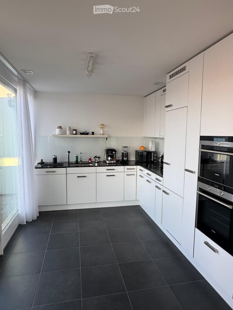 Modern kitchen with white cabinets, black countertops, and stainless steel appliances, including an oven and microwave. The space features a large window with white curtains and a shelf with decorative items.