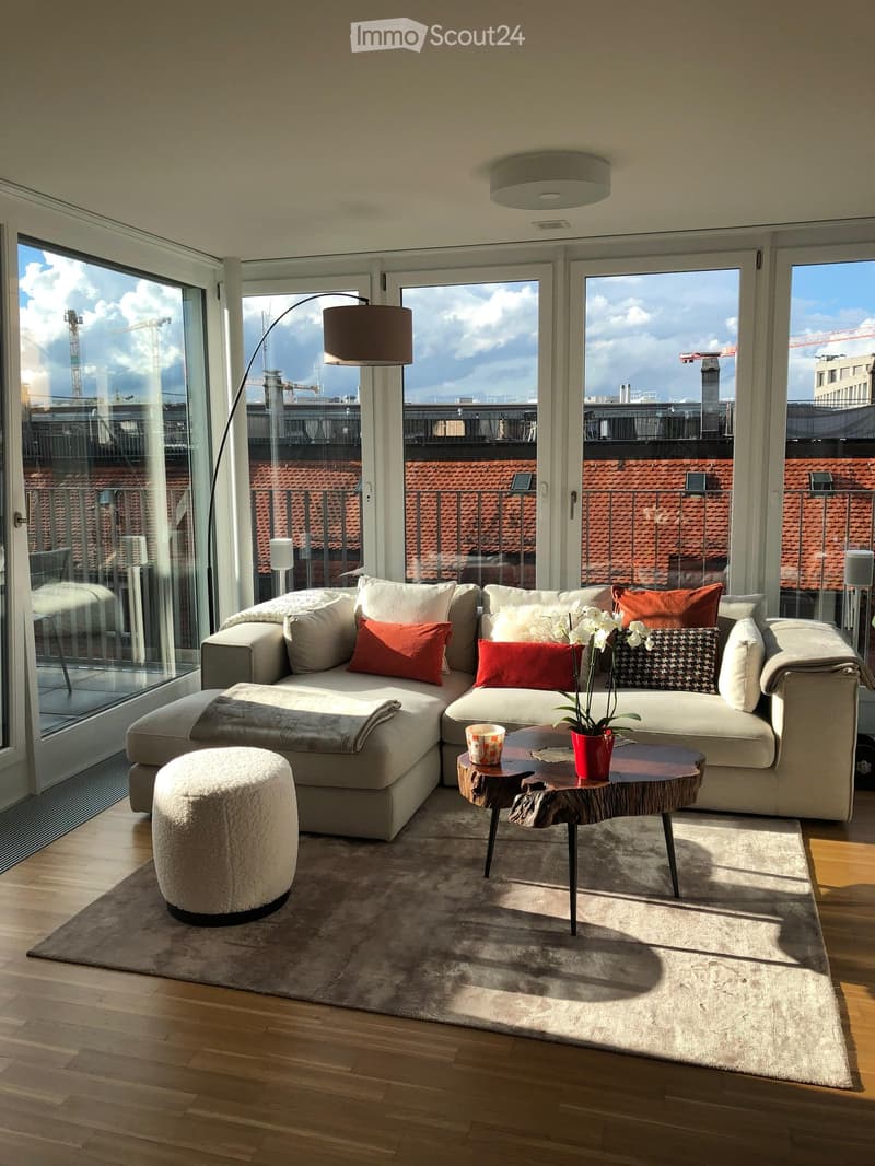 Modern living room with large windows offering a city view, featuring a sectional sofa, coffee table, floor lamp, and a decorative flower pot.