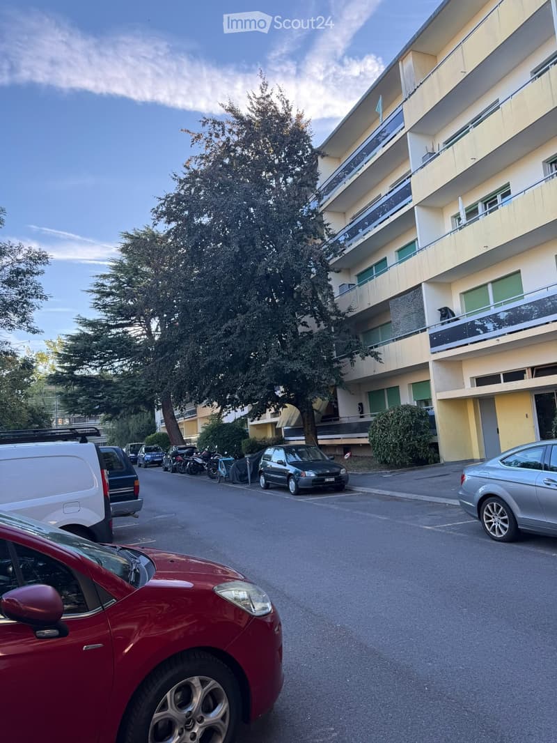 Multi-story residential building with balconies, surrounded by parked cars and trees.