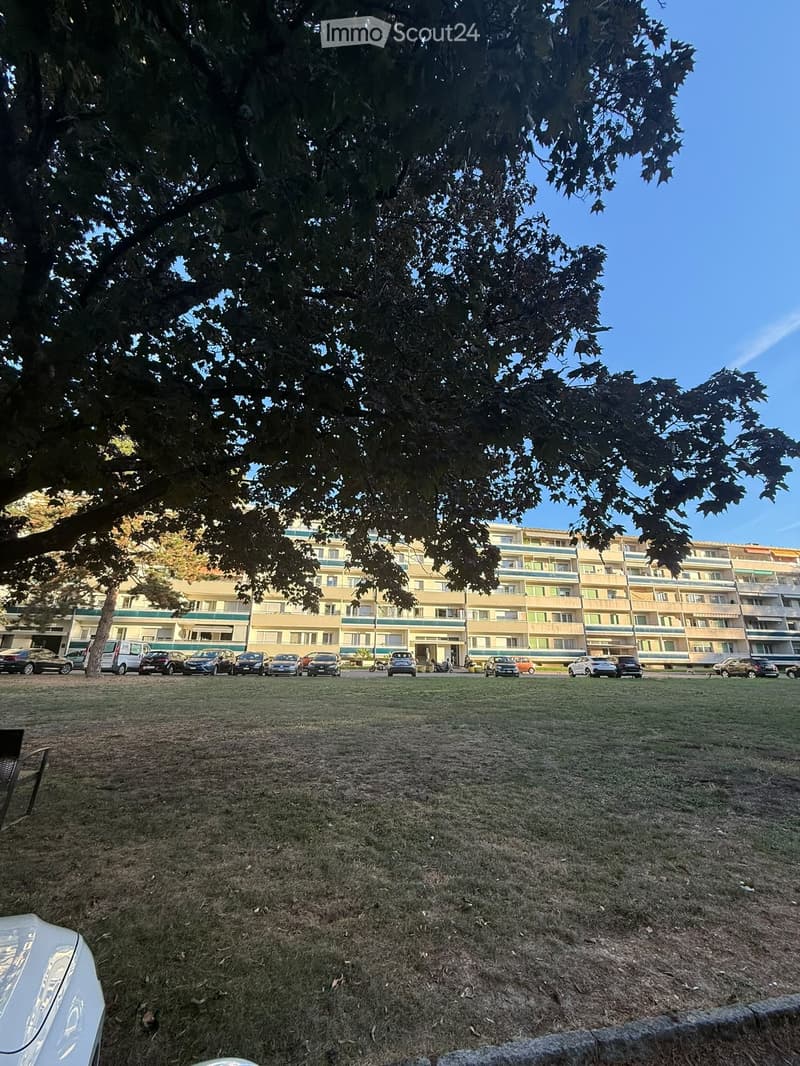 Multi-story residential building with balconies, surrounded by a grassy area with parked cars.