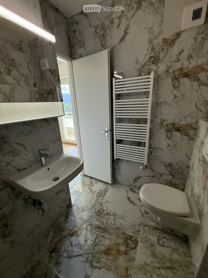 Modern bathroom with marble walls and floor, featuring a sink, toilet, and a white radiator. An open door leads to a balcony area.