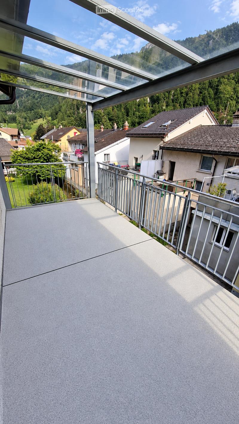 Balcony with a modern glass roof, metal railing, and a view of a residential area with houses and green hills.