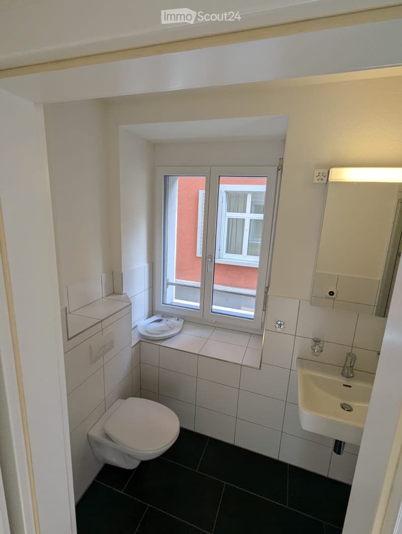 Compact bathroom with white tiled walls, black tiled floor, toilet, sink, and small window offering a view of the exterior building.