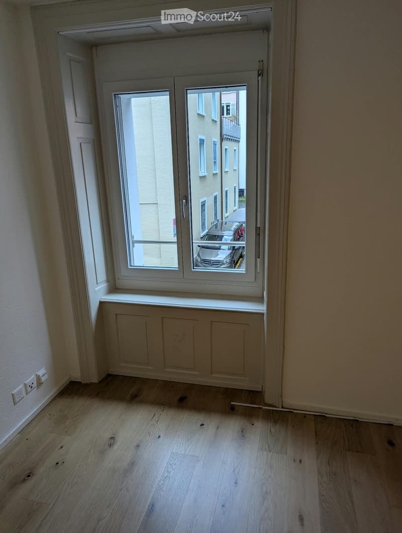 Empty room with light wooden flooring, white walls, and a large window with a view of the street and parked cars.