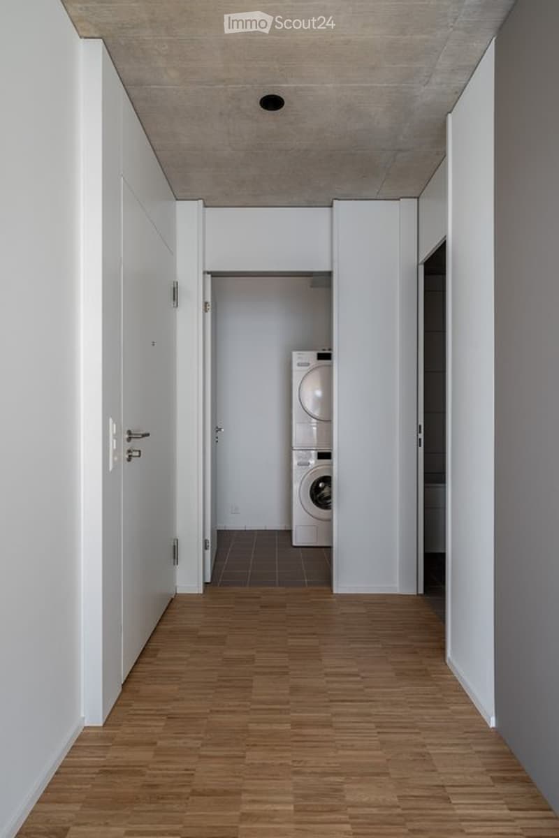 Clean, modern hallway with wooden flooring, white walls, and a private washing machine visible in an adjacent room.