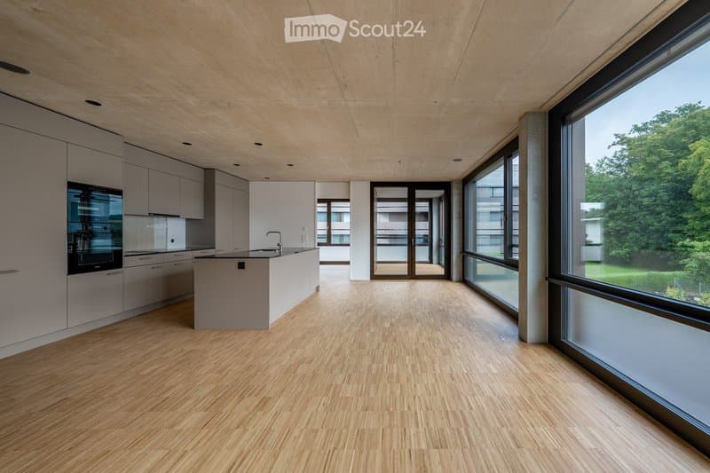 Open-plan kitchen and living area with light wood flooring, white cabinetry, and large windows offering a view of greenery. The space is unfurnished, with only built-in cabinetry and a kitchen island visible.