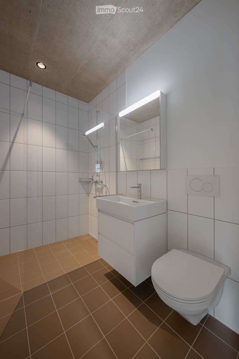 Modern bathroom with white tiles, a white sink with a chrome faucet, a toilet, a large mirror with built-in lighting, and a shower area with a glass enclosure.