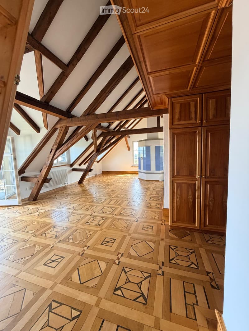 An attic space with exposed wooden beams, a patterned wooden floor, and white walls. The room features wooden doors and windows, with natural light streaming in from the windows.
