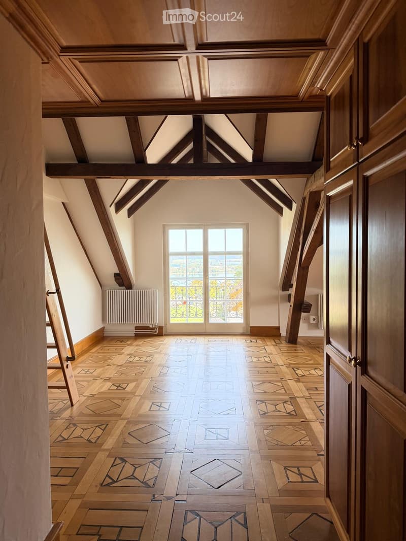 An empty attic space with exposed wooden beams, a large window with decorative ironwork, and a patterned wooden floor.