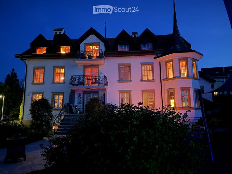 Large, elegant two-story house with white walls, multiple windows, and a steeply pitched roof featuring dormers and a prominent spire. The house is illuminated from within, with warm lighting visible through the windows, and is surrounded by well-maintain