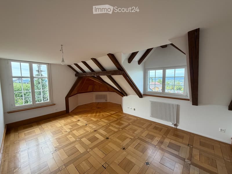 Empty attic room with wooden floors, exposed wooden beams, large windows, and radiators on the walls.