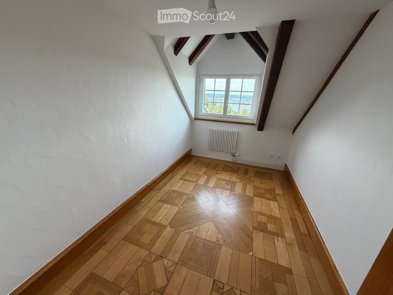 Empty attic room with wooden herringbone flooring, white walls, exposed wooden beams, and a single window offering a scenic view.