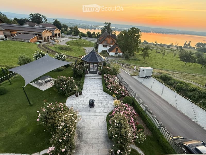 Aerial view of a house with a garden, a gazebo, a winding road, a small lake, and a sunset in the background.