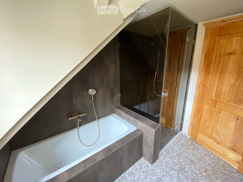 Corner bathtub, shower area with glass door, wooden door, tiled walls and floor, separate bathtub and shower area