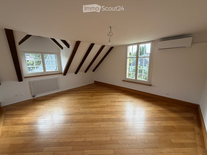 Empty attic room with wooden flooring, white walls, exposed wooden beams, and large windows allowing natural light.