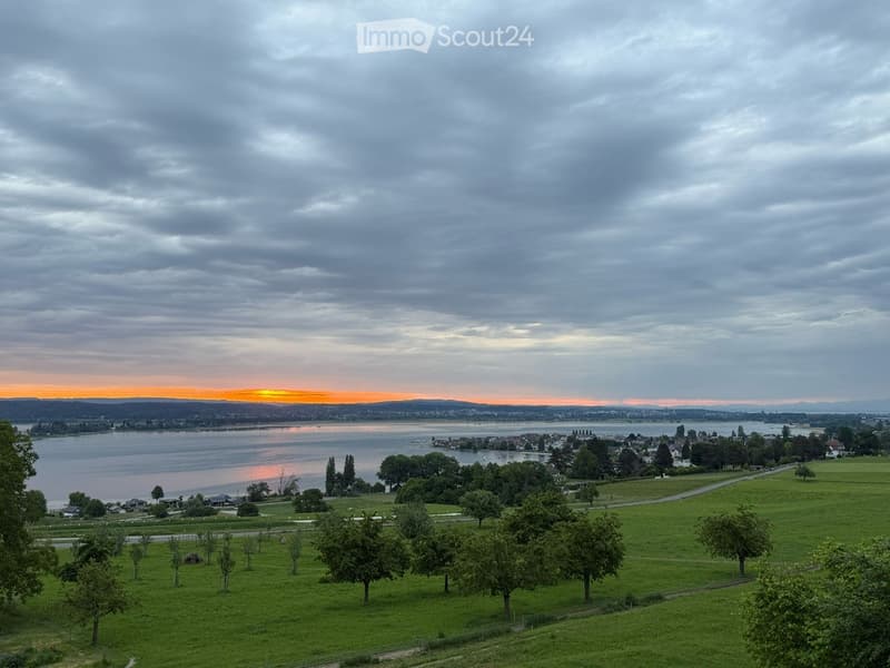 Wide view of a landscape with a body of water, green hills, and a sunset in the background.