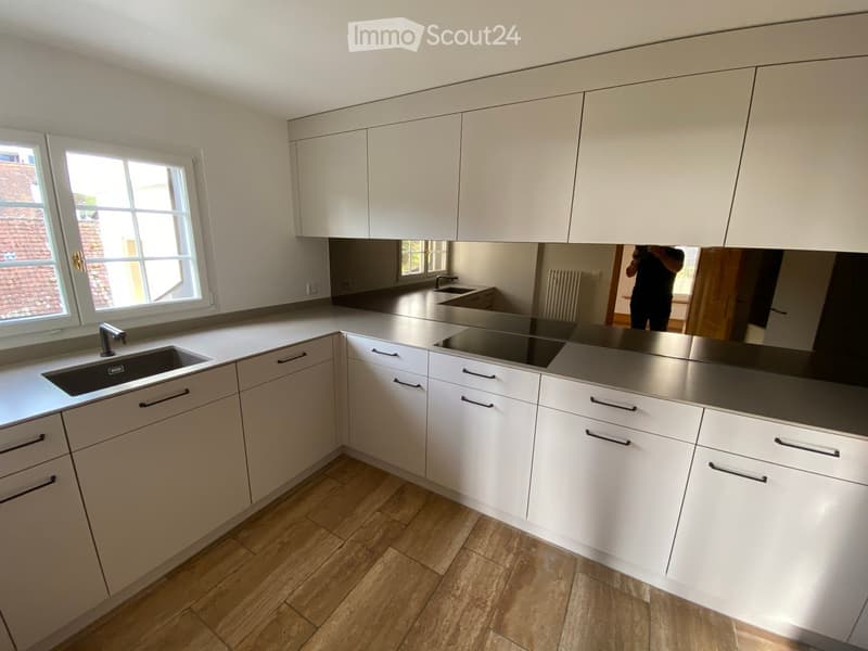 Modern kitchen with white cabinets, stainless steel countertops, large windows, wooden flooring, single sink, black induction stove, mirror with reflection, small room at back