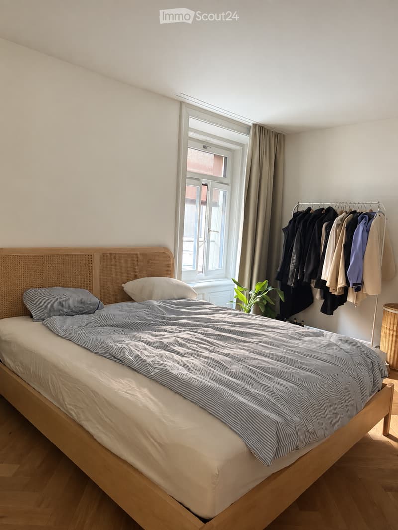 A well-lit bedroom with a wooden bed, striped bedding, a wicker headboard, a coat rack with jackets, a small plant, and a wicker basket near the window.