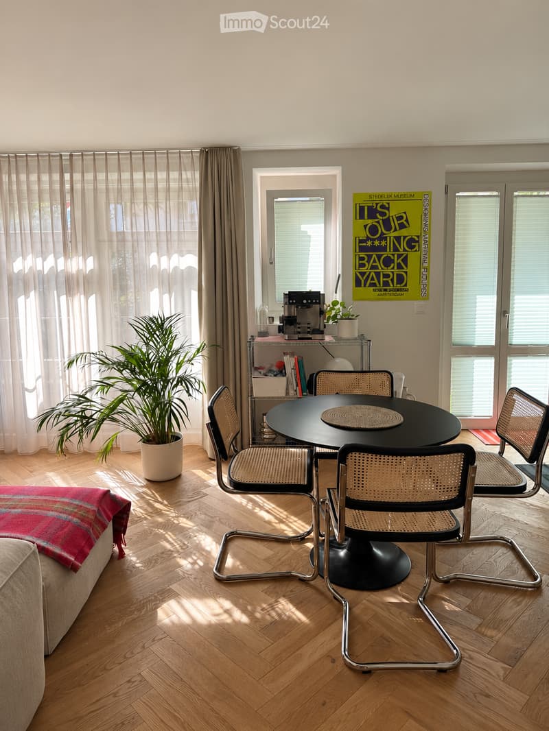 Bright living room with wooden herringbone flooring, large windows with sheer curtains, a round black dining table with four chairs, a sofa with a red and blue blanket, a potted plant, and a coffee machine on a shelf.