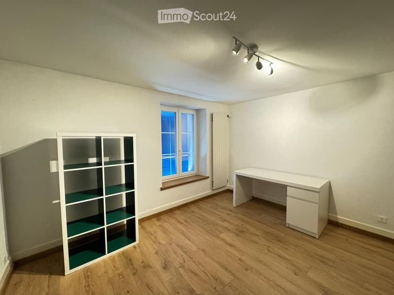 A small, unfurnished room with wooden flooring, a white shelf, and a white desk near a window.