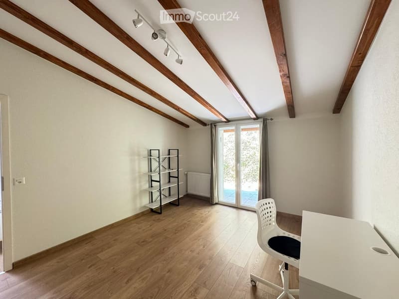 Bright, airy room with wooden flooring, exposed wooden beams, and large windows. Features a white desk, a white chair, and a black shelving unit.