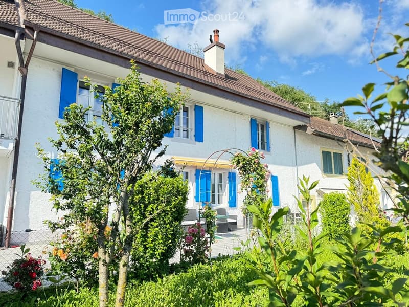 Two-story white house with blue shutters, surrounded by lush greenery and a small patio with outdoor furniture.