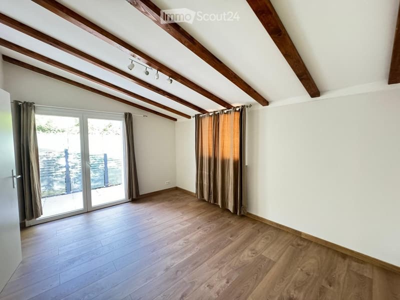 Empty attic room with wooden beams, wooden flooring, large sliding glass door with curtains, and recessed lighting.