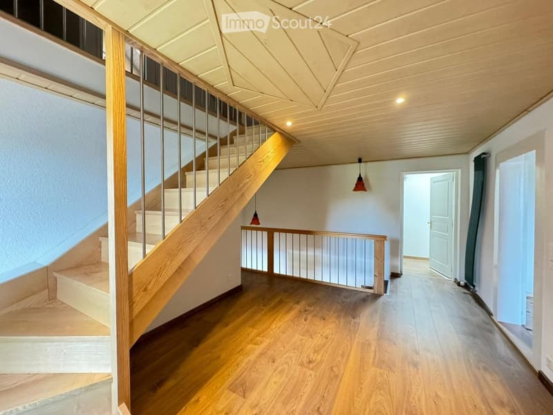 Spacious hallway with wooden flooring, a staircase with wooden steps and metal railing, and pendant lights hanging from the ceiling.