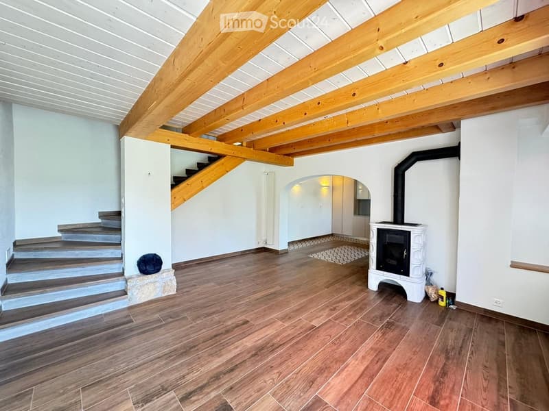 Spacious room with wooden flooring, exposed wooden beams, a staircase leading to an upper level, and a white fireplace with a black chimney.
