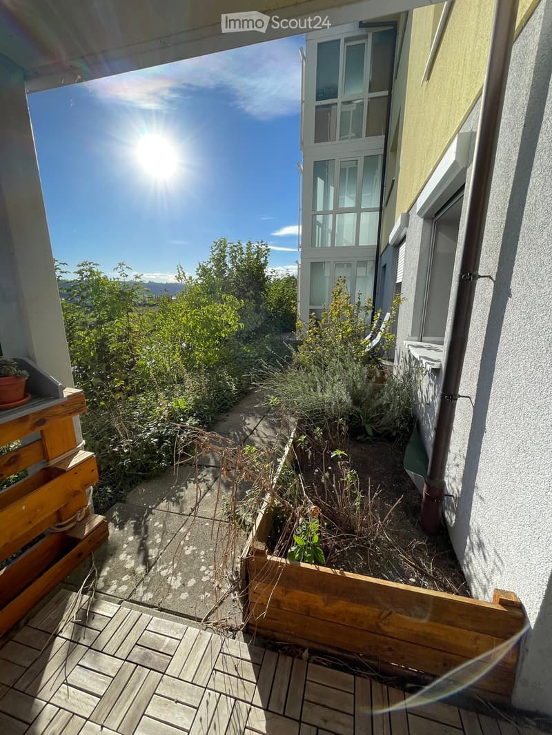 Balcony with wooden decking, small garden area with plants and a wooden planter box, overlooking a building with large windows.
