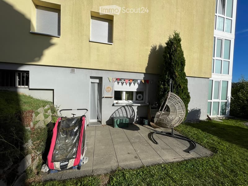 Garden area in front of a two-story house with a concrete patio, a hanging egg chair, a stroller, and a small green table.