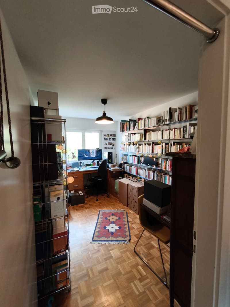 A well-lit study or home office with wooden flooring, a desk with a laptop and chair, bookshelves filled with books, and a colorful rug on the floor.