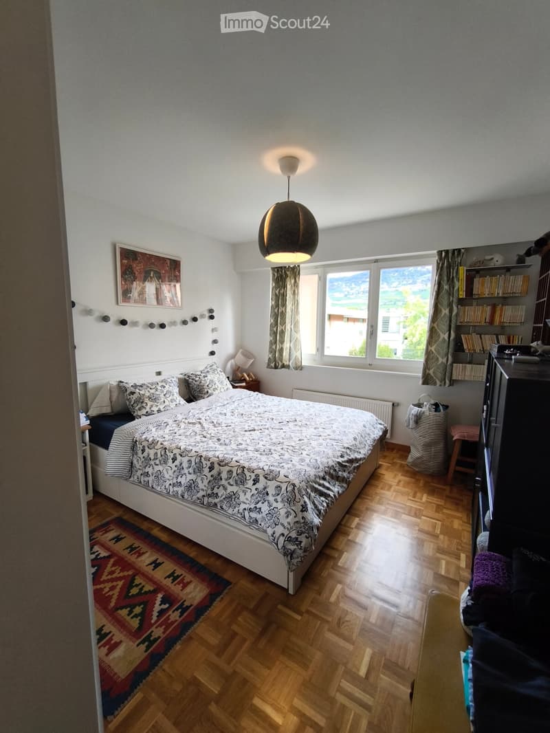 Cozy bedroom with wooden flooring, a large bed with patterned bedding, a nightstand with a lamp, a bookshelf, and a view through large windows.