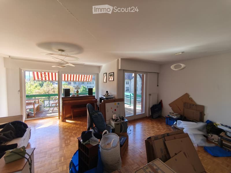 Living room with wooden flooring, large windows with striped blinds, piano, sofa, table, chairs, and various items including boxes and bags.