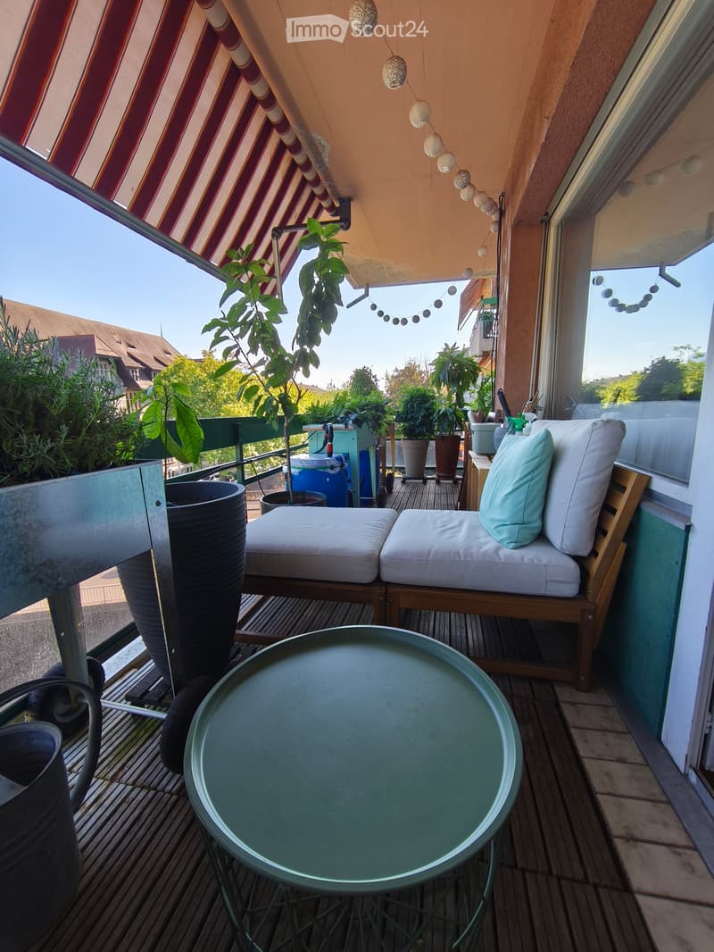 Balcony with wooden decking, white cushioned seating, round table, and several potted plants.