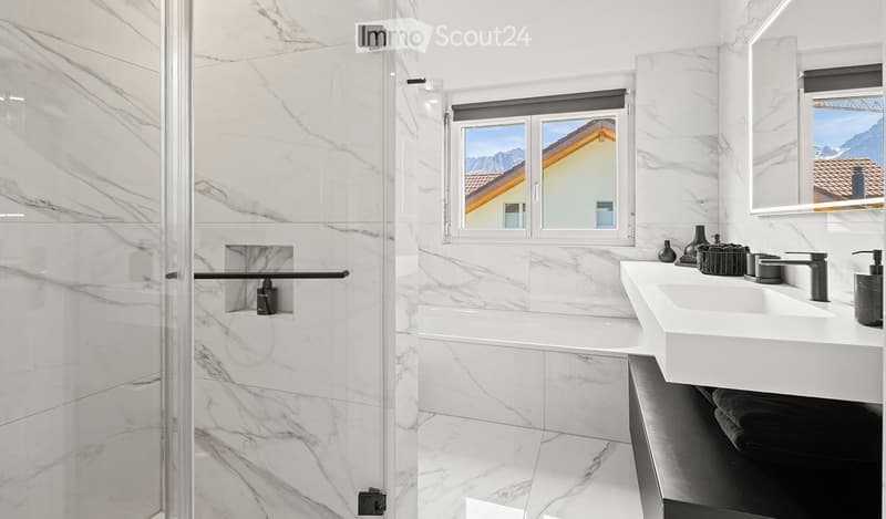 Modern bathroom with marble tiles, a bathtub, a double sink, and a glass-enclosed shower. The room features large windows with a scenic mountain view.