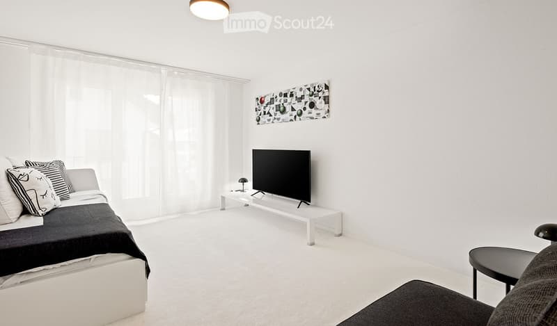 Modern, minimalist room with white walls, a bed, a sofa, a TV on a stand, and a small table with a lamp.