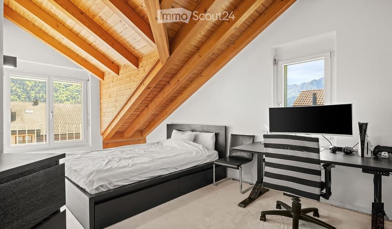 Modern bedroom with a wooden slanted ceiling, large windows offering a scenic view, a neatly made bed, a desk with a screen, and a comfortable chair.