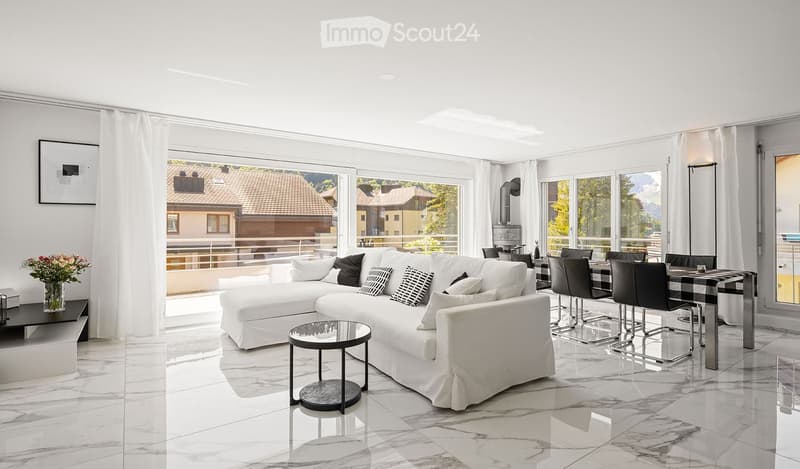 Modern living room with white sofa, coffee table, dining area, large windows offering a scenic mountain view, and decorative elements like a vase with flowers and a picture frame on the wall.