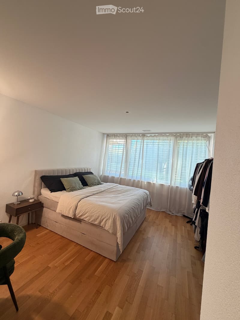 A modern bedroom with a large bed, nightstand, chair, and window with curtains. The room features wooden flooring and white walls.